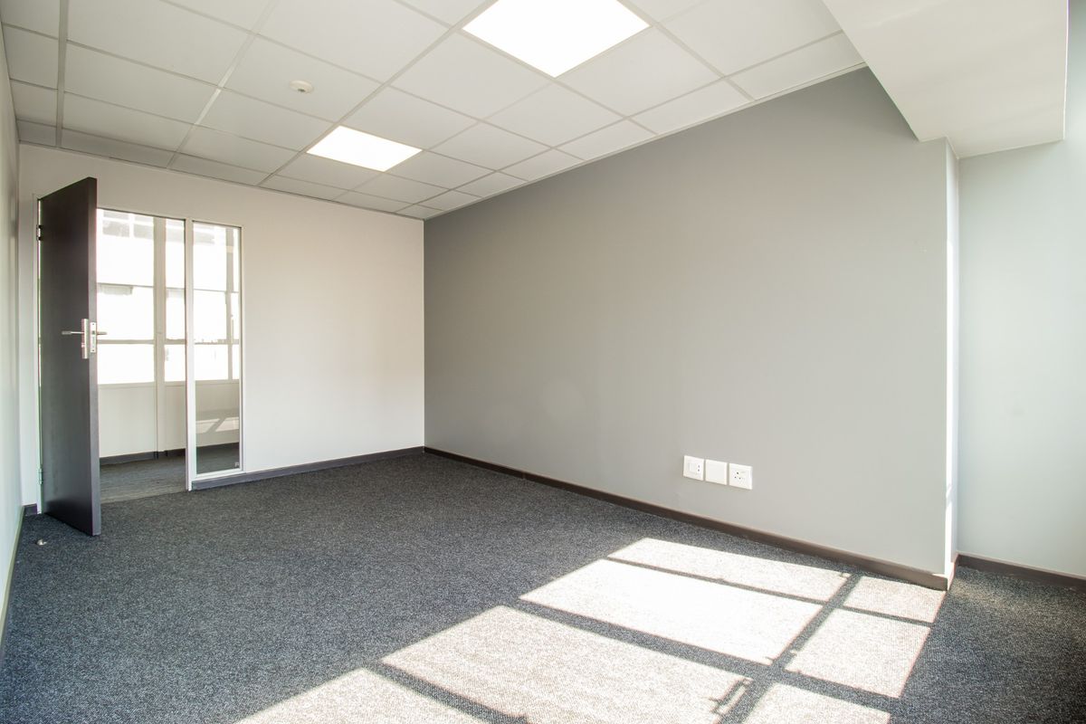 A clean, vacant office space featuring grey carpeting, neutral walls, a suspended ceiling with recessed lighting, and a glass-paneled entrance door.