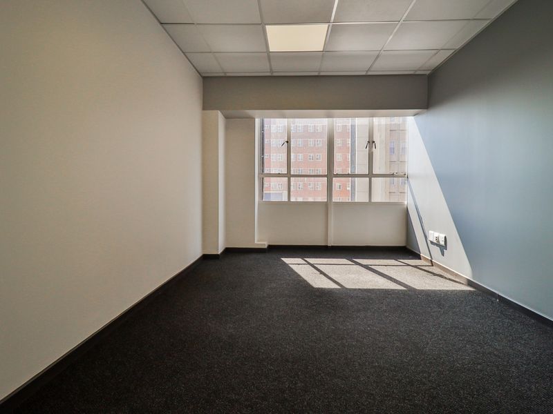 A clean, empty office space featuring neutral walls, dark carpet flooring, a suspended ceiling with recessed lighting, and a large window providing natural light.