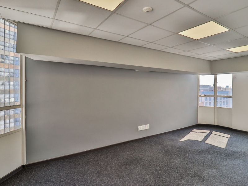 A bright, empty commercial office space featuring grey carpeting, a suspended ceiling with recessed lighting, and large windows providing natural light.