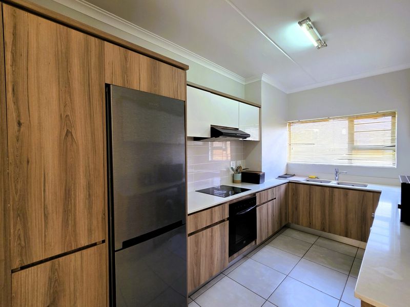 Modern kitchen featuring wood-grain cabinetry, white countertops, integrated electric hob and oven, tiled backsplash, and a double sink with natural light.