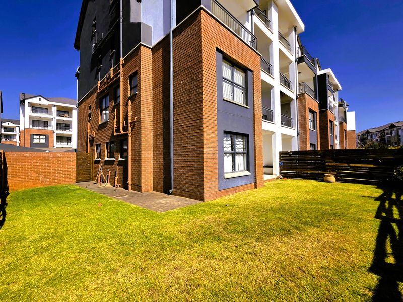 Rear view of a modern apartment building featuring a brick facade, private balconies, and a well-maintained garden area with lawn and fencing.