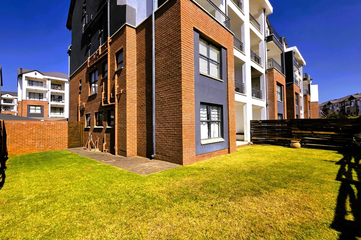 Rear view of a modern apartment building featuring a brick facade, private balconies, and a well-maintained garden area with lawn and fencing.