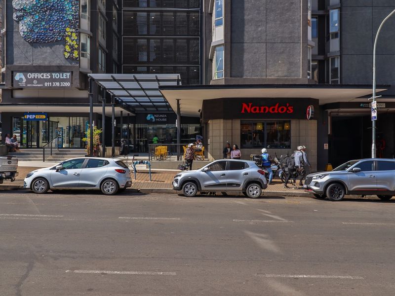 Street-level view of a commercial building facade featuring a Nando's storefront, covered entrance canopy, and paved sidewalk access in an urban setting.