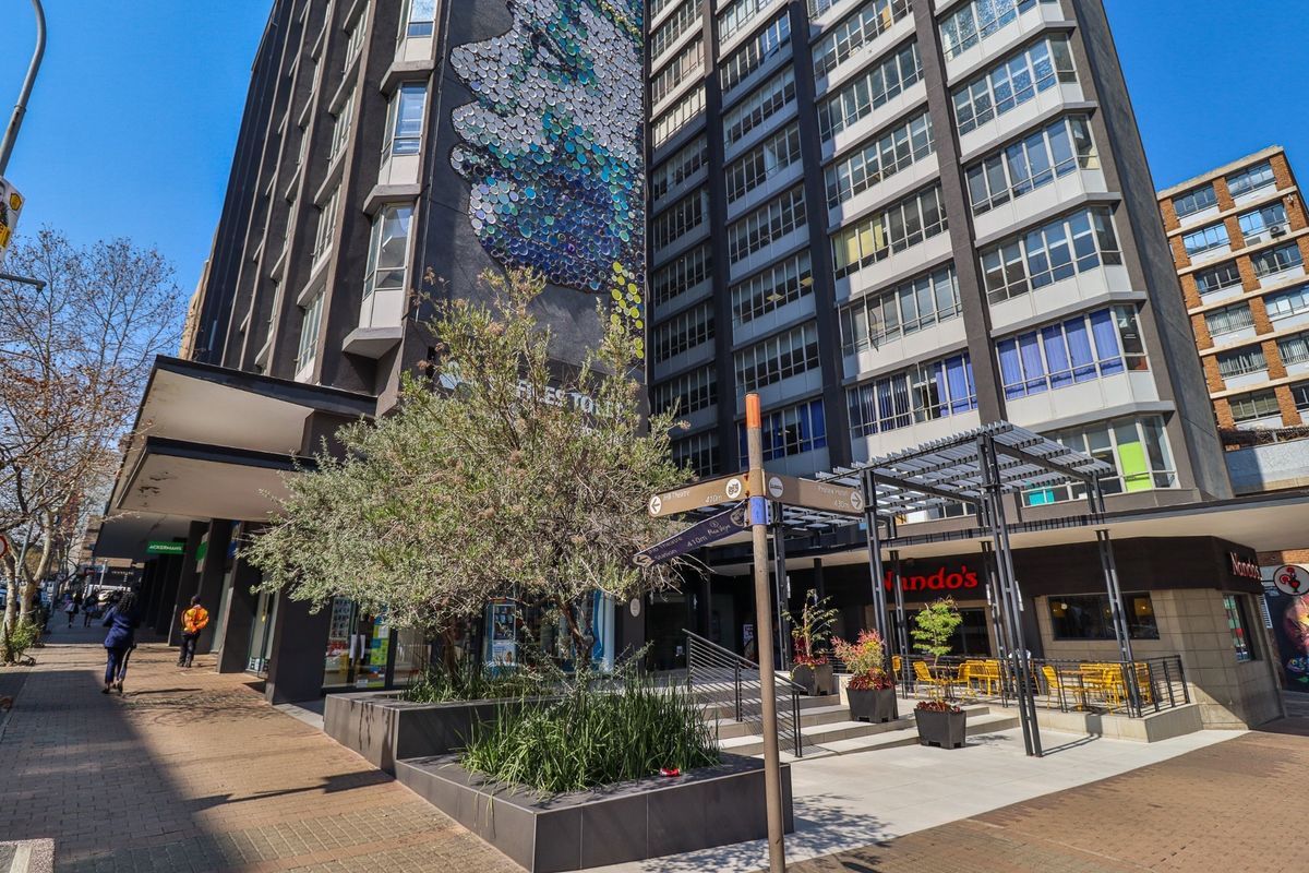 Exterior view of a multi-story commercial building featuring a prominent wall mural, paved pedestrian plaza, and integrated outdoor seating area with a metal pergola.