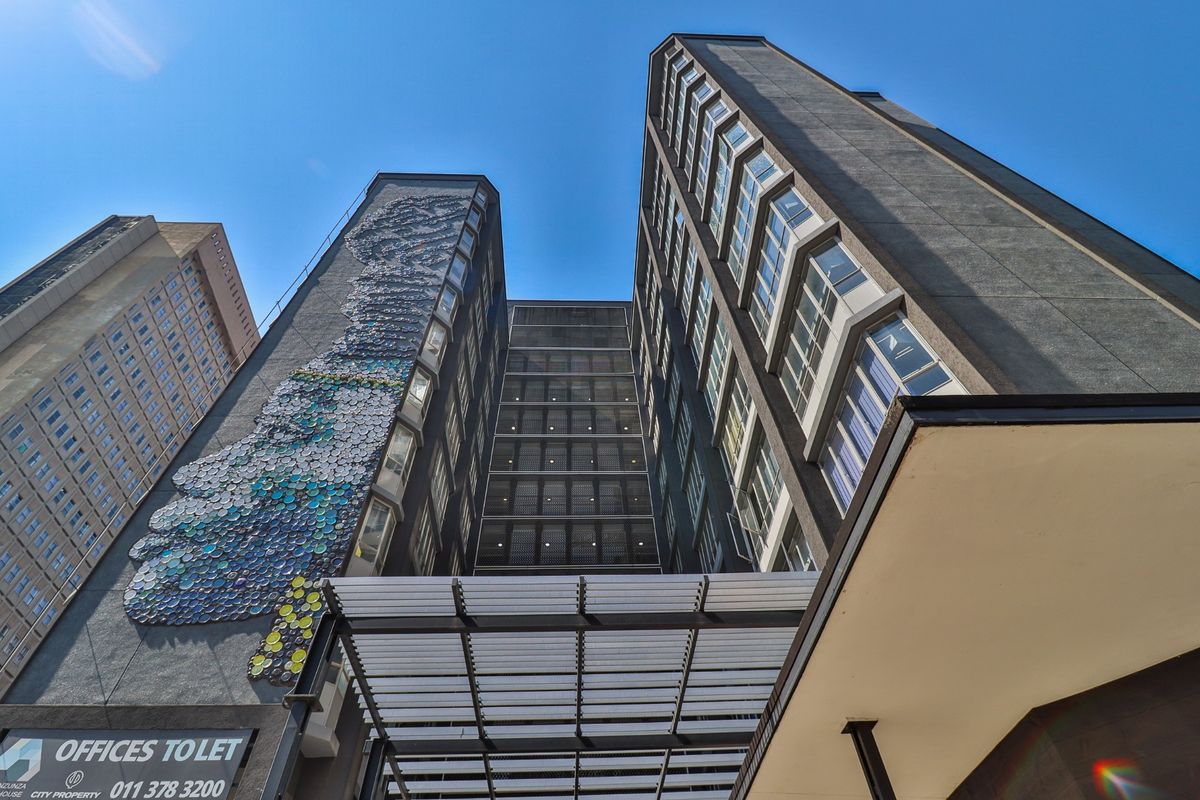 Low-angle view of a modern commercial high-rise building featuring a prominent artistic wall mural, metal entrance canopy, and extensive glass window facade.