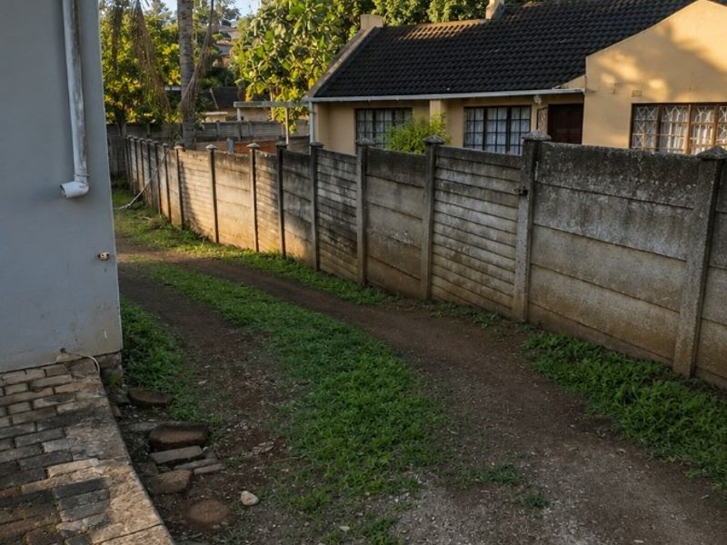 A view of the rear exterior area featuring a precast concrete boundary wall, a paved walkway, and an unpaved driveway leading toward the neighboring property.
