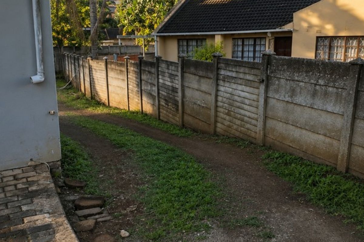 A view of the rear exterior area featuring a precast concrete boundary wall, a paved walkway, and an unpaved driveway leading toward the neighboring property.