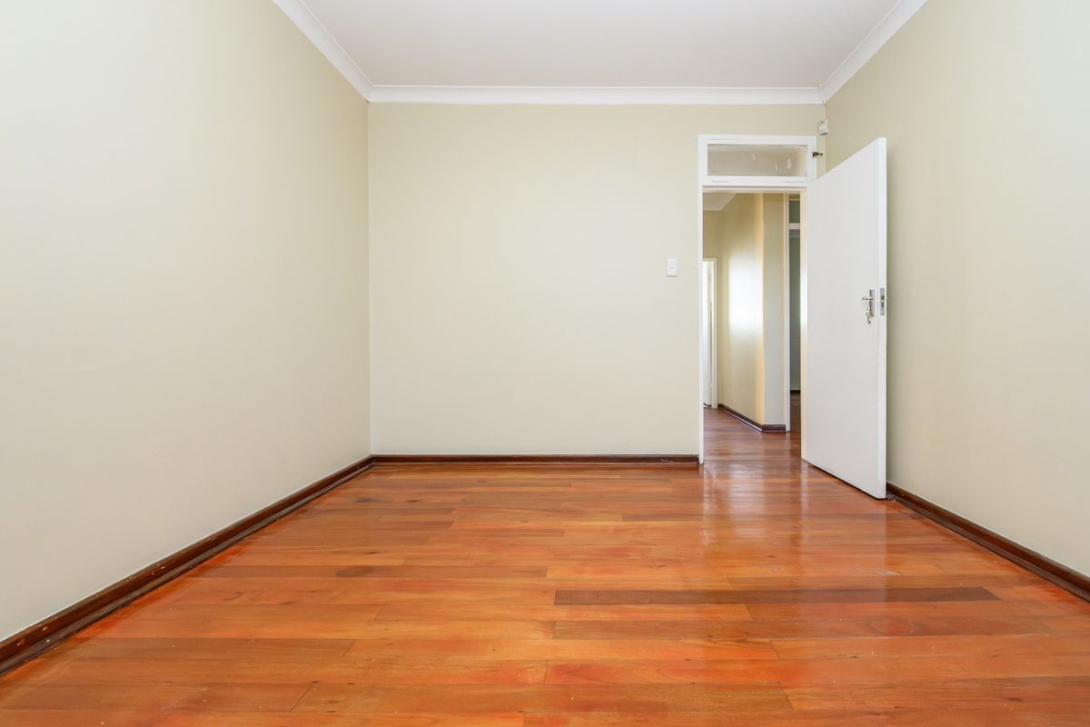 A bright, empty bedroom featuring polished hardwood flooring, neutral walls, and a classic internal door with a transom window.