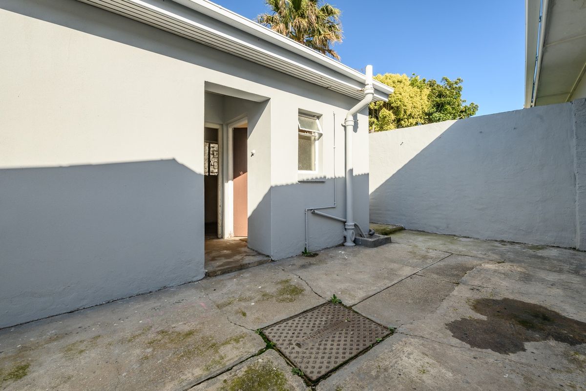 A rear exterior view of the property featuring a concrete paved courtyard, boundary wall, and external drainage infrastructure.