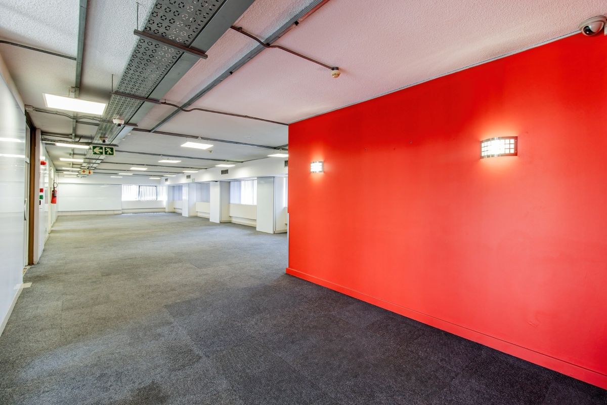 An open-plan commercial office space featuring exposed industrial cable trays, recessed lighting, and a prominent red feature wall.
