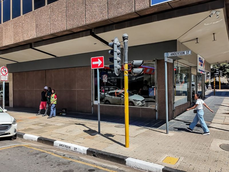 Street-level view of a commercial building corner featuring large display windows, paved sidewalk access, and prominent street signage at the intersection.