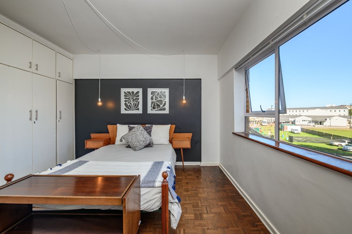 A bright bedroom featuring classic parquet flooring, ample built-in cupboard storage, and a large window offering natural light and exterior views.