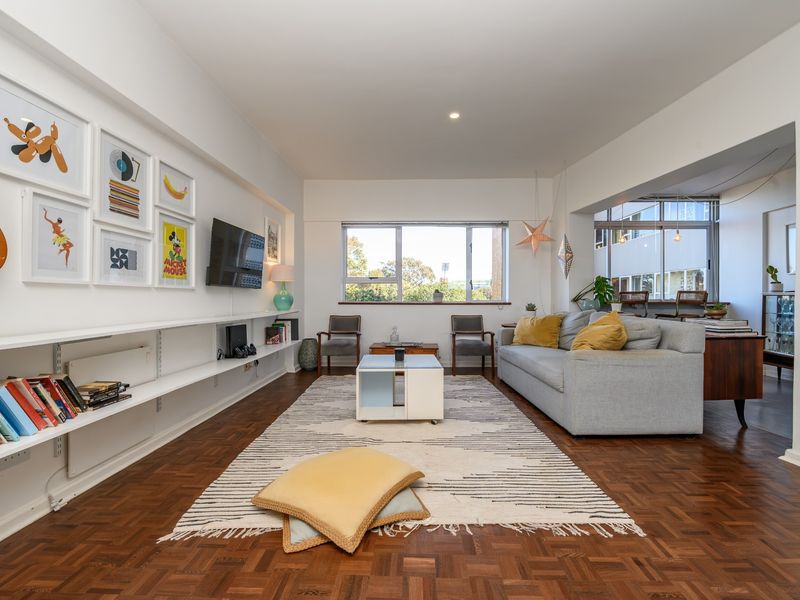 A bright and spacious living room featuring classic parquet flooring, built-in wall shelving, and large windows that provide ample natural light.