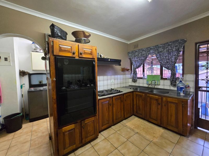 A functional kitchen featuring wooden cabinetry, built-in oven, hob, extractor fan, and a double sink with tiled flooring and backsplash.