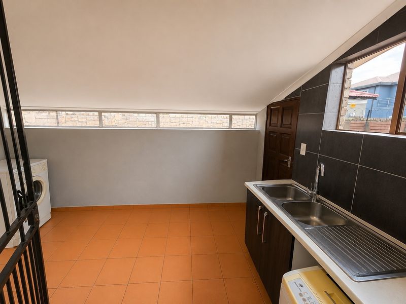A functional laundry room featuring a double stainless steel sink, dark tiled splashback, built-in cabinetry, and orange floor tiles under a sloped ceiling.