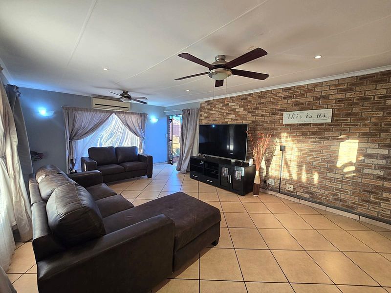 Spacious living room featuring an exposed brick accent wall, tiled flooring, ceiling fans, and a wall-mounted air conditioning unit.