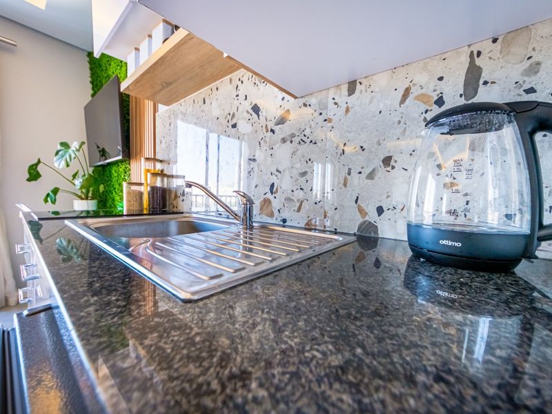 Modern kitchen area featuring a dark granite countertop, stainless steel sink, and a stylish terrazzo splashback with integrated cabinetry and shelving.