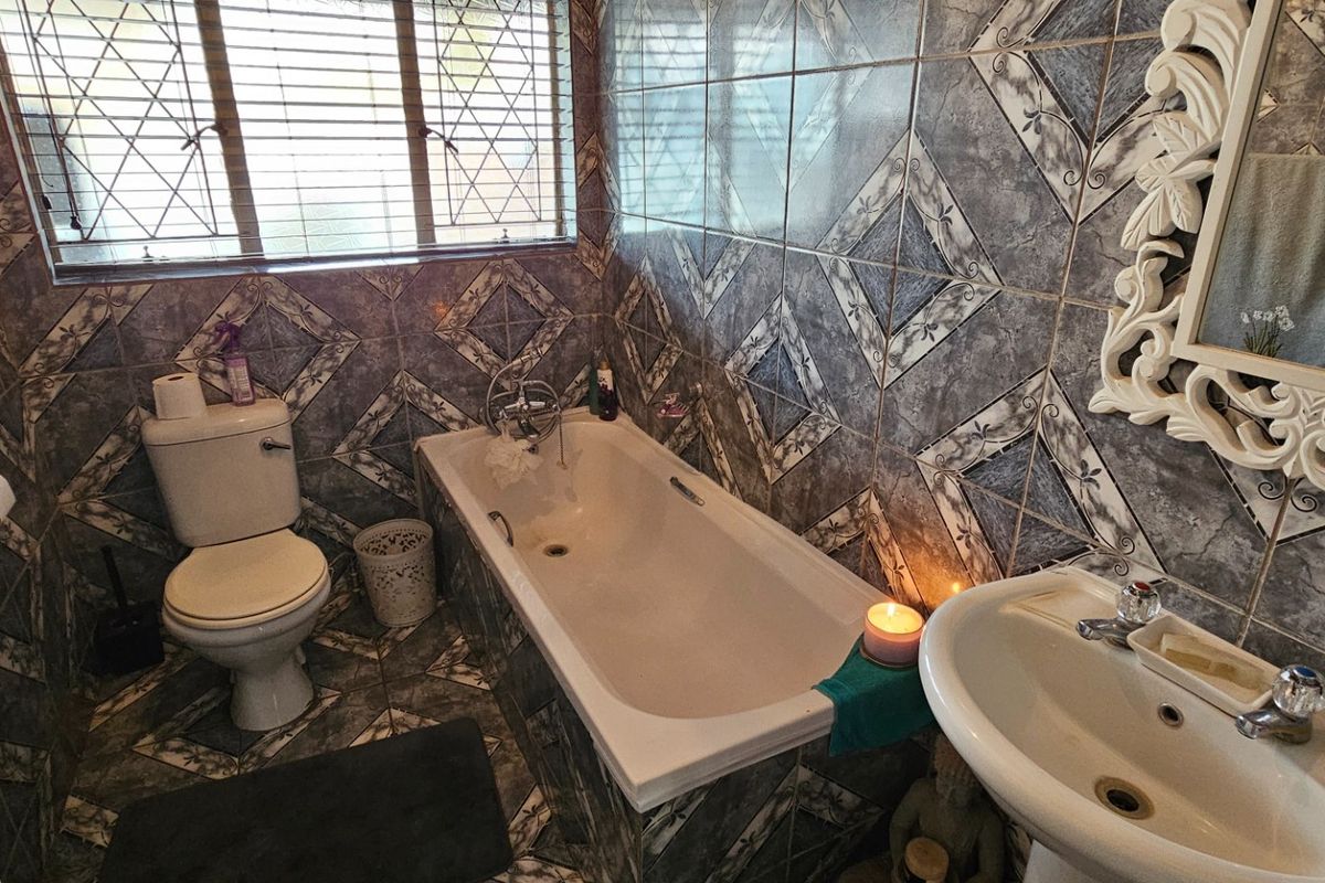 A functional bathroom featuring a bathtub, pedestal basin, and toilet, finished with patterned wall and floor tiles and a window with security bars.