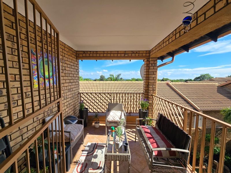 A covered balcony featuring exposed brick walls, tiled flooring, and metal balustrades, offering an elevated outdoor space with views of the surrounding townhouse complex.