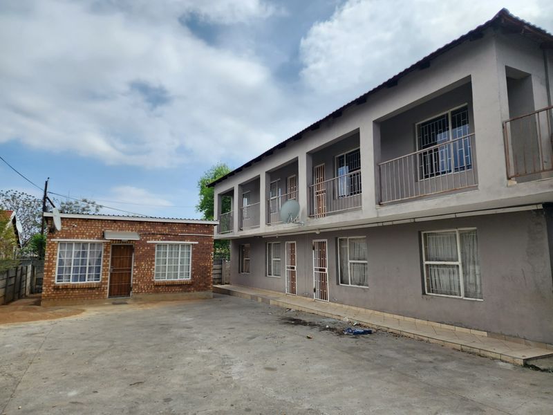 Exterior view of a multi-story apartment building with a paved courtyard, featuring brick and plastered facades, security gates, and balcony access.