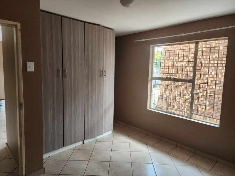 A clean, empty bedroom featuring neutral tiled flooring, built-in wooden cupboards, and a window fitted with security bars.