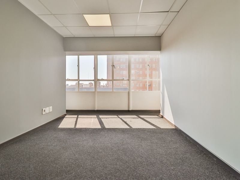 A bright, empty office space featuring neutral walls, carpeted flooring, a suspended ceiling with recessed lighting, and large windows providing natural light.