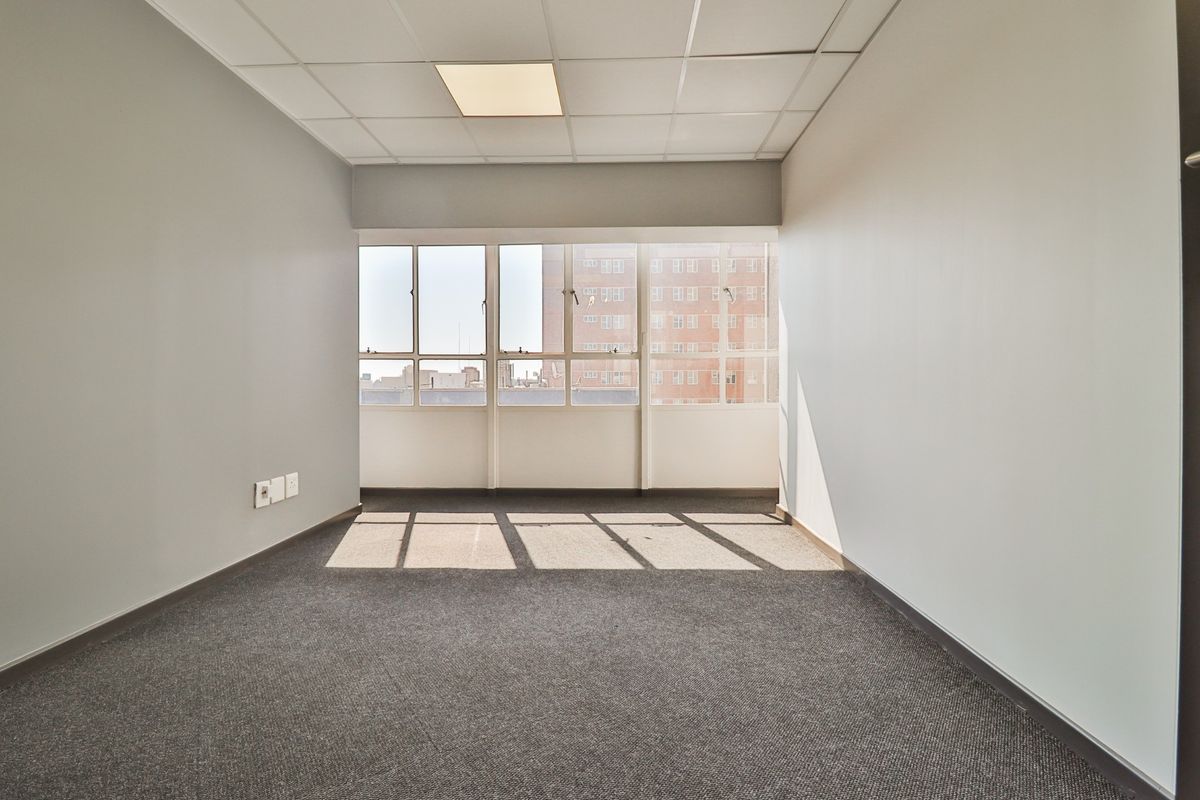 A bright, empty office space featuring neutral walls, carpeted flooring, a suspended ceiling with recessed lighting, and large windows providing natural light.