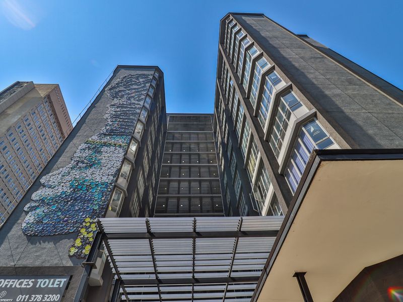 Low-angle view of a modern commercial high-rise building featuring a prominent artistic wall mural, metal entrance canopy, and extensive glass window facade.