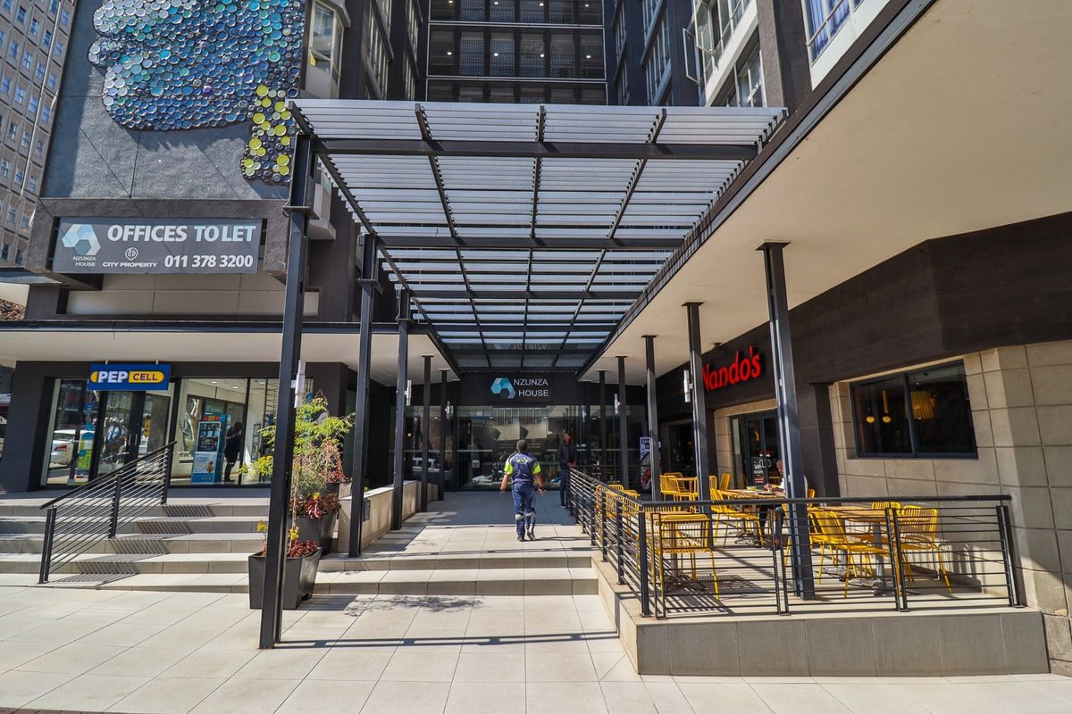 Modern commercial building entrance featuring a metal pergola, paved walkway, and glass doors, providing access to office and retail spaces.
