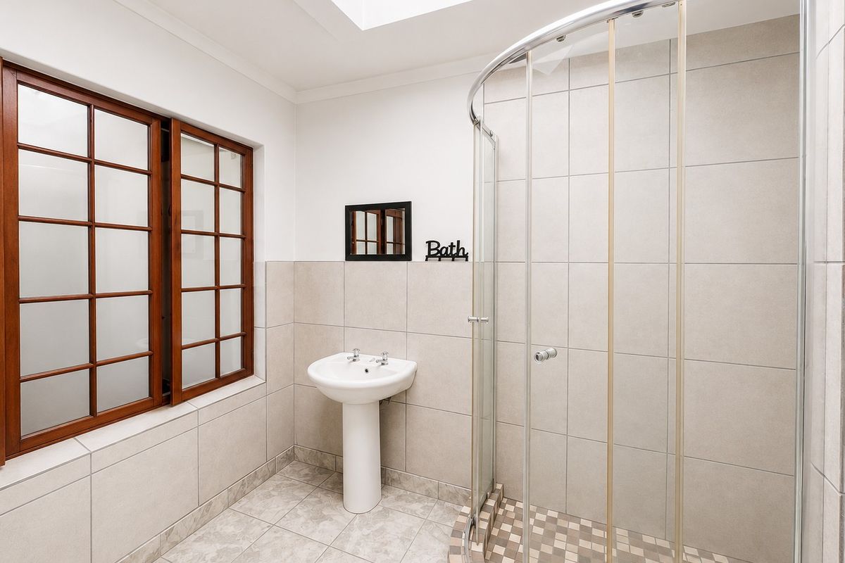 A bright bathroom featuring a pedestal basin, a curved shower enclosure, and a skylight, finished with neutral wall and floor tiling.