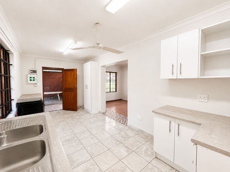 A clean, functional kitchen featuring white cabinetry, laminate countertops, double stainless steel sink, tiled flooring, and a ceiling fan, leading to an adjacent room.