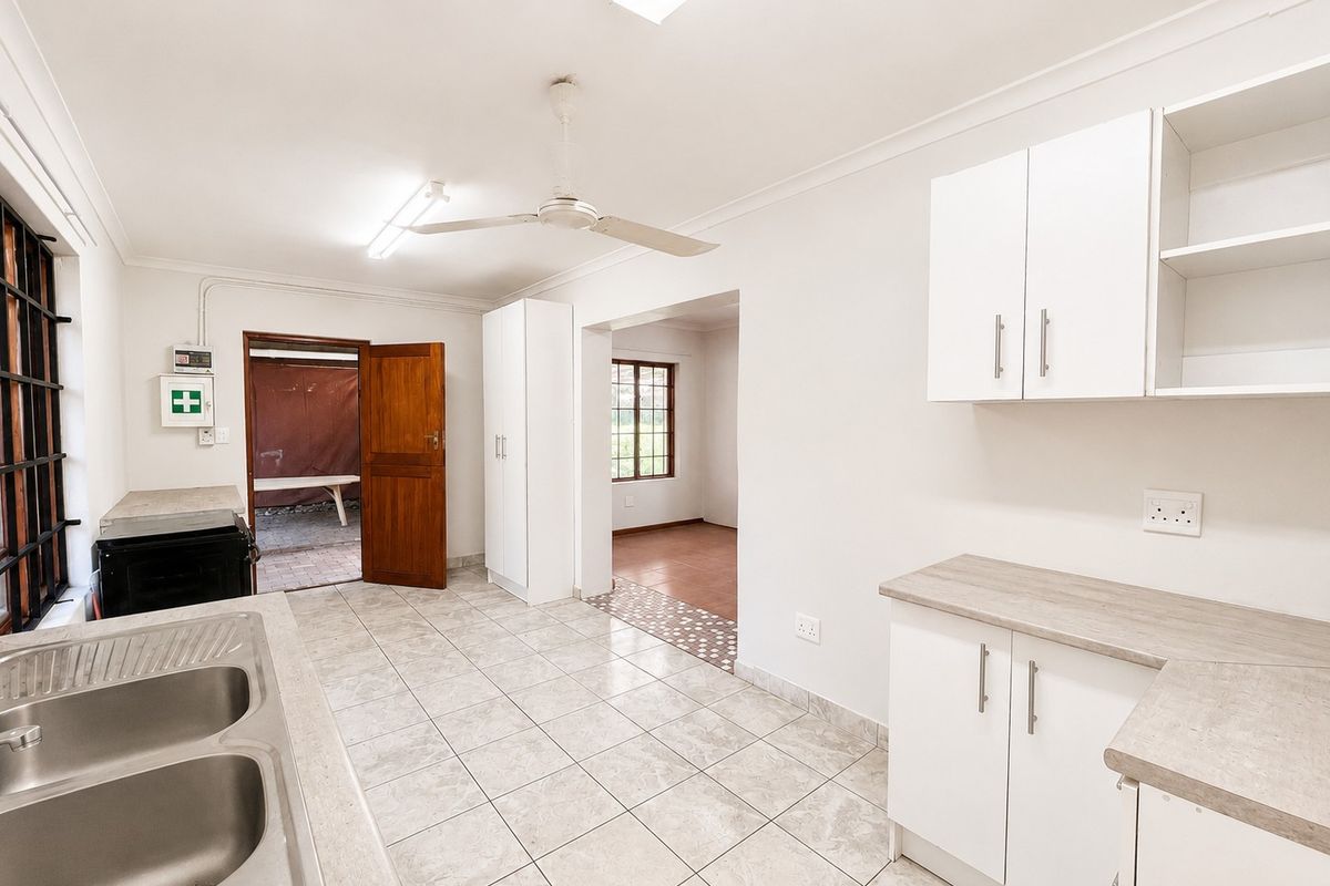 A clean, functional kitchen featuring white cabinetry, laminate countertops, double stainless steel sink, tiled flooring, and a ceiling fan, leading to an adjacent room.