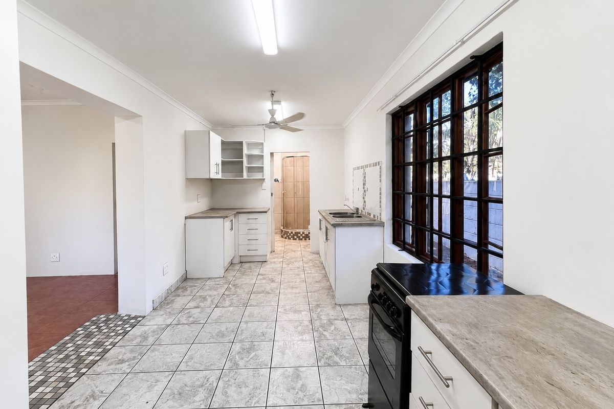 A functional, empty kitchen featuring tiled flooring, laminate countertops, built-in cabinetry, and a large timber-framed window providing natural light.