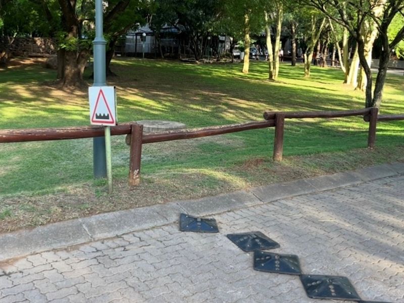 A street-level view of the property frontage featuring a paved road with speed bumps, a wooden boundary fence, and mature trees in the background.