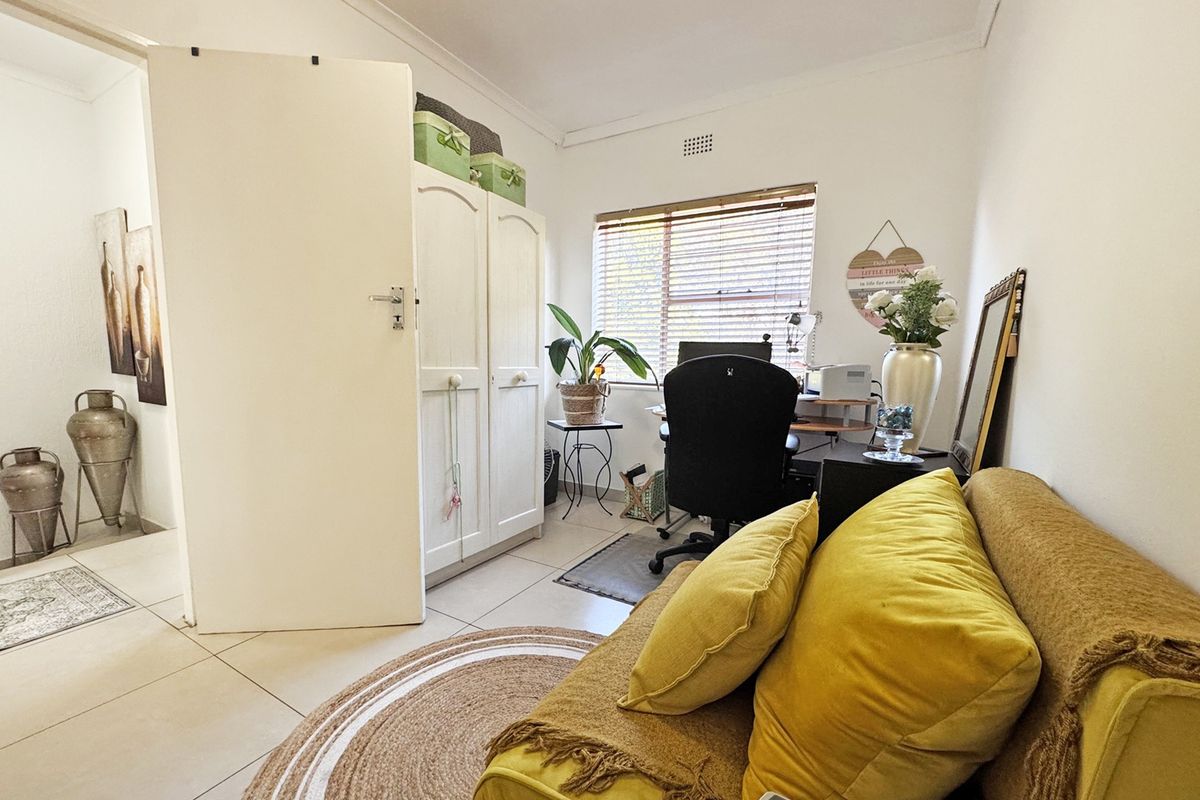 A functional bedroom featuring tiled flooring, a built-in wardrobe, and a window with venetian blinds, currently utilized as a home office space.