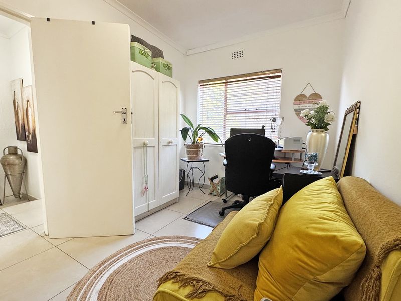 A functional bedroom featuring tiled flooring, a built-in wardrobe, and a window with venetian blinds, currently utilized as a home office space.