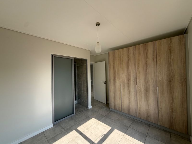 A clean, modern bedroom featuring neutral tiled flooring, a large built-in wood-finish wardrobe, and a sliding door leading to an en-suite bathroom.