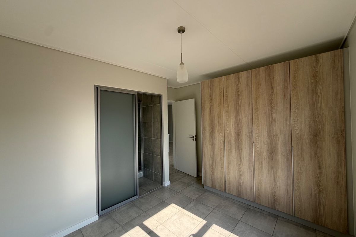 A clean, modern bedroom featuring neutral tiled flooring, a large built-in wood-finish wardrobe, and a sliding door leading to an en-suite bathroom.
