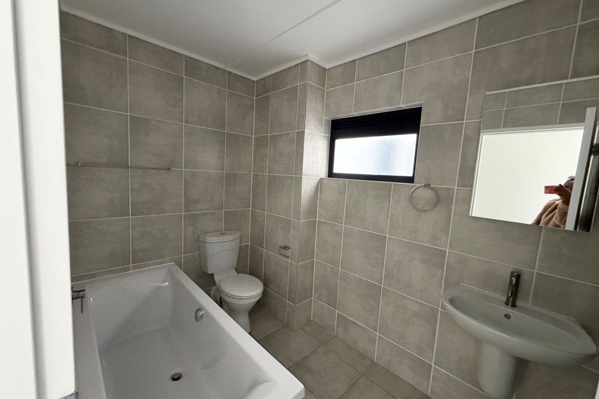 A clean, modern bathroom featuring neutral grey wall and floor tiles, a white bathtub, pedestal basin, and a functional layout.