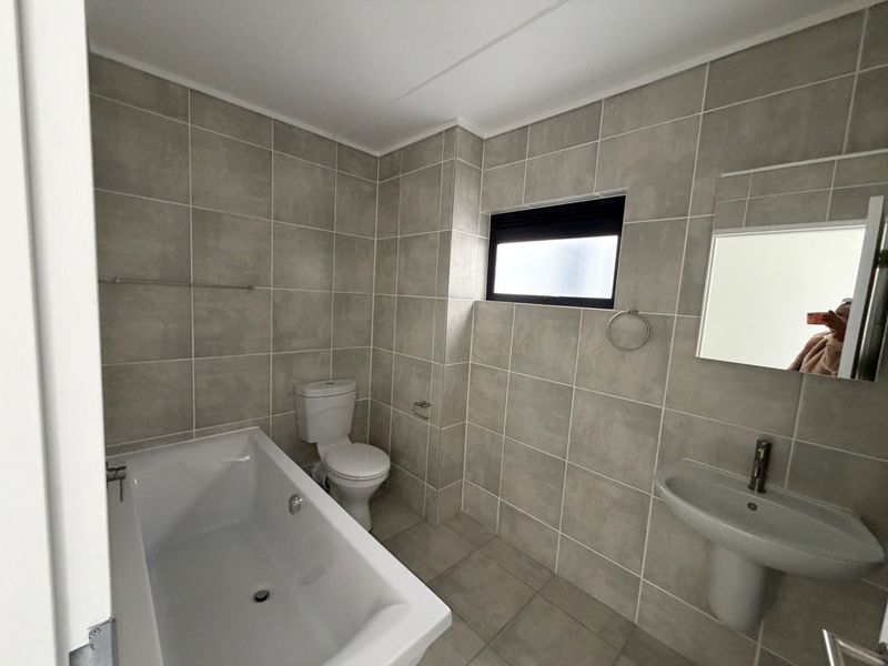 A clean, modern bathroom featuring neutral grey wall and floor tiles, a white bathtub, pedestal basin, and a functional layout.