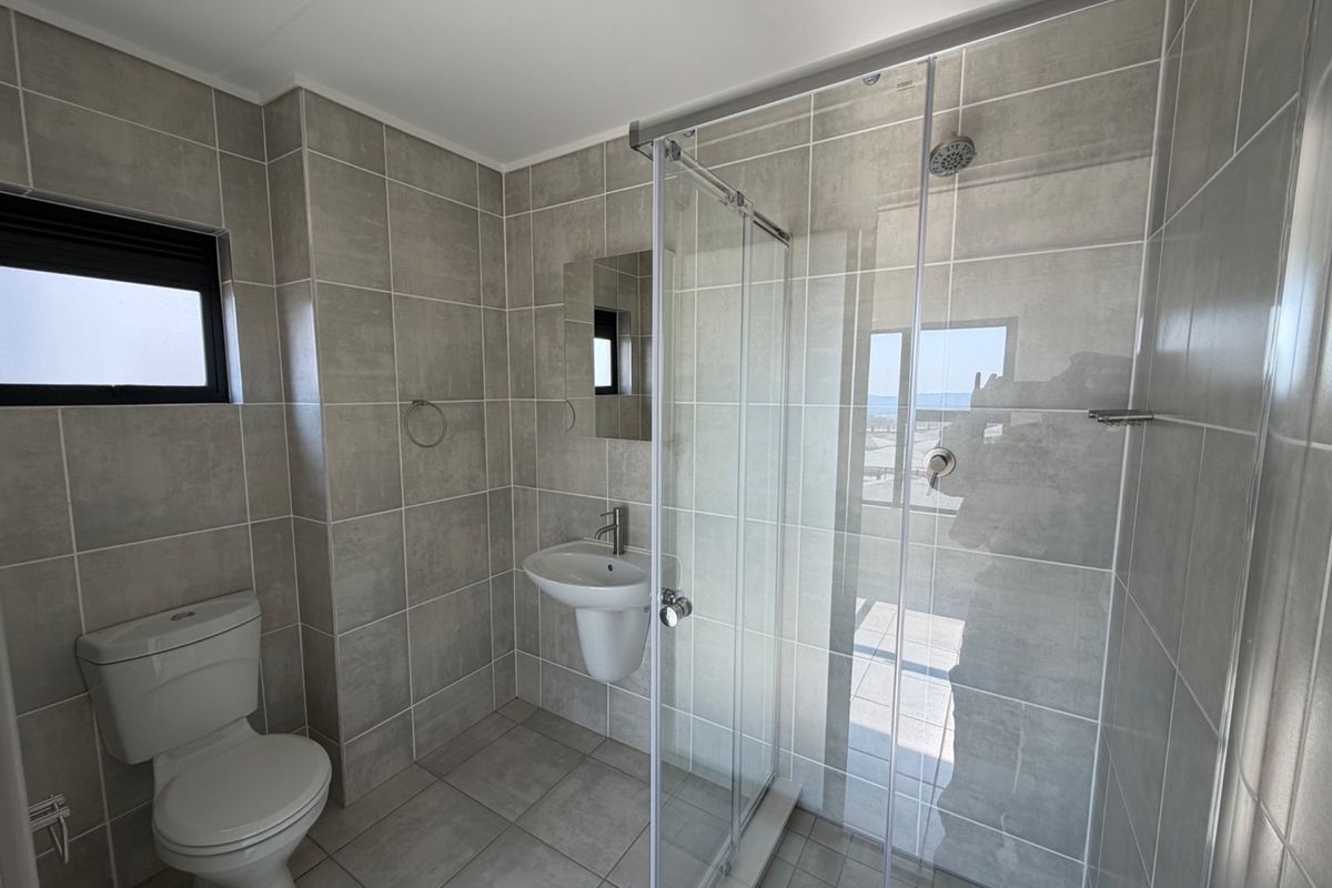 A modern, clean bathroom featuring neutral ceramic tiling, a glass-enclosed shower, a pedestal washbasin, and a standard toilet.