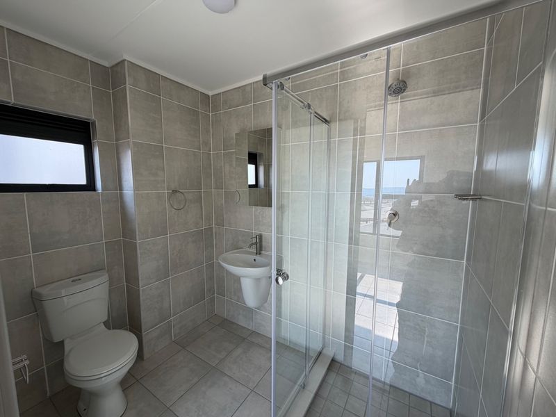 A modern, clean bathroom featuring neutral ceramic tiling, a glass-enclosed shower, a pedestal washbasin, and a standard toilet.