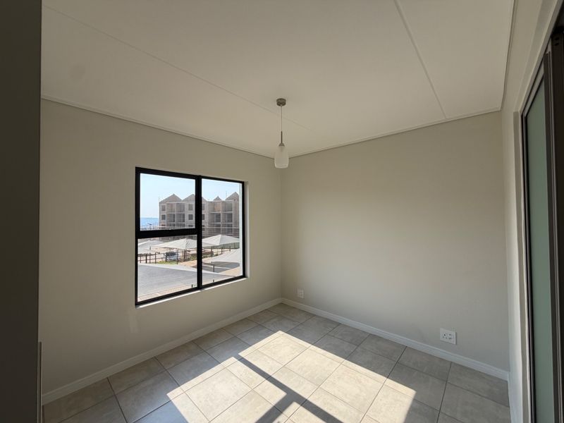 A bright, empty bedroom featuring neutral tiled flooring, a modern black aluminium window frame, and a simple pendant light fixture.