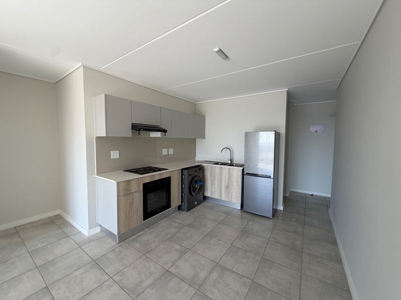Modern kitchen featuring light wood-tone cabinetry, built-in oven and hob, stainless steel sink, and neutral tiled flooring in an open-plan layout.