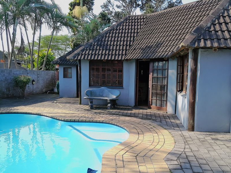 A backyard pool area featuring a paved deck, swimming pool, and a single-story dwelling with a tiled roof and timber-framed windows.