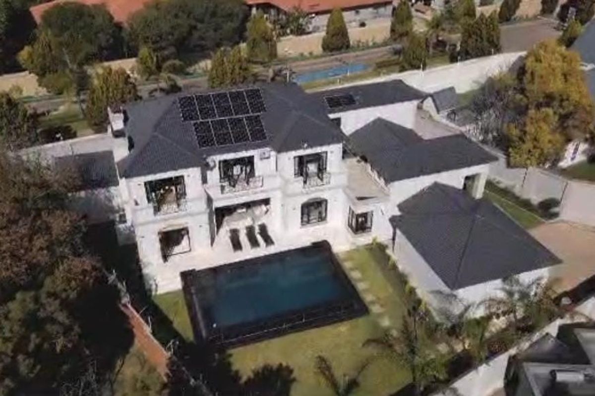 Aerial view of a large luxury residence featuring a dark tiled roof, integrated solar panels, a private swimming pool, and landscaped garden areas.