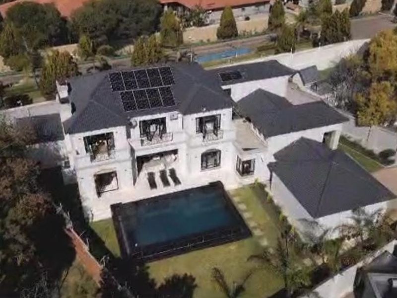 Aerial view of a large luxury residence featuring a dark tiled roof, integrated solar panels, a private swimming pool, and landscaped garden areas.