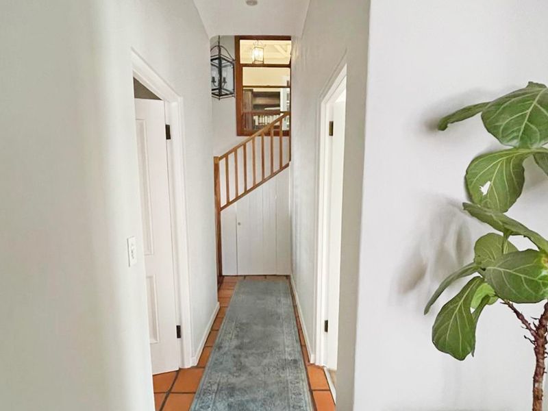 A bright hallway featuring terracotta floor tiles, a wooden staircase balustrade, and access to multiple rooms via internal doors.