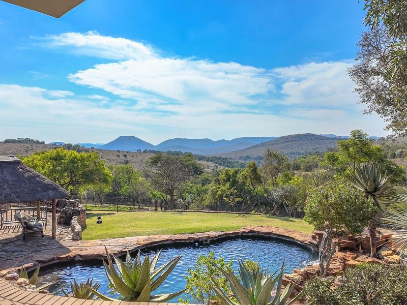 Scenic outdoor view featuring a natural-style swimming pool, thatched lapa, and expansive landscaped garden overlooking rolling hills and mountains.