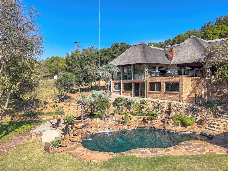 Rear view of a farm-style residence featuring a thatched roof, brick facade, and a natural rock pool set within a landscaped garden.